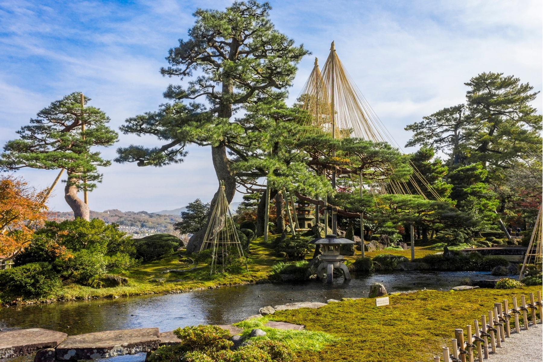 A wide panoramic view of Kenroku-en Garden in Kanazawa, featuring ancient pine trees with traditional Yukitsuri rope supports, a stone Kotoji-toro lantern by the pond, and a blue sky over the Ishikawa landscape.