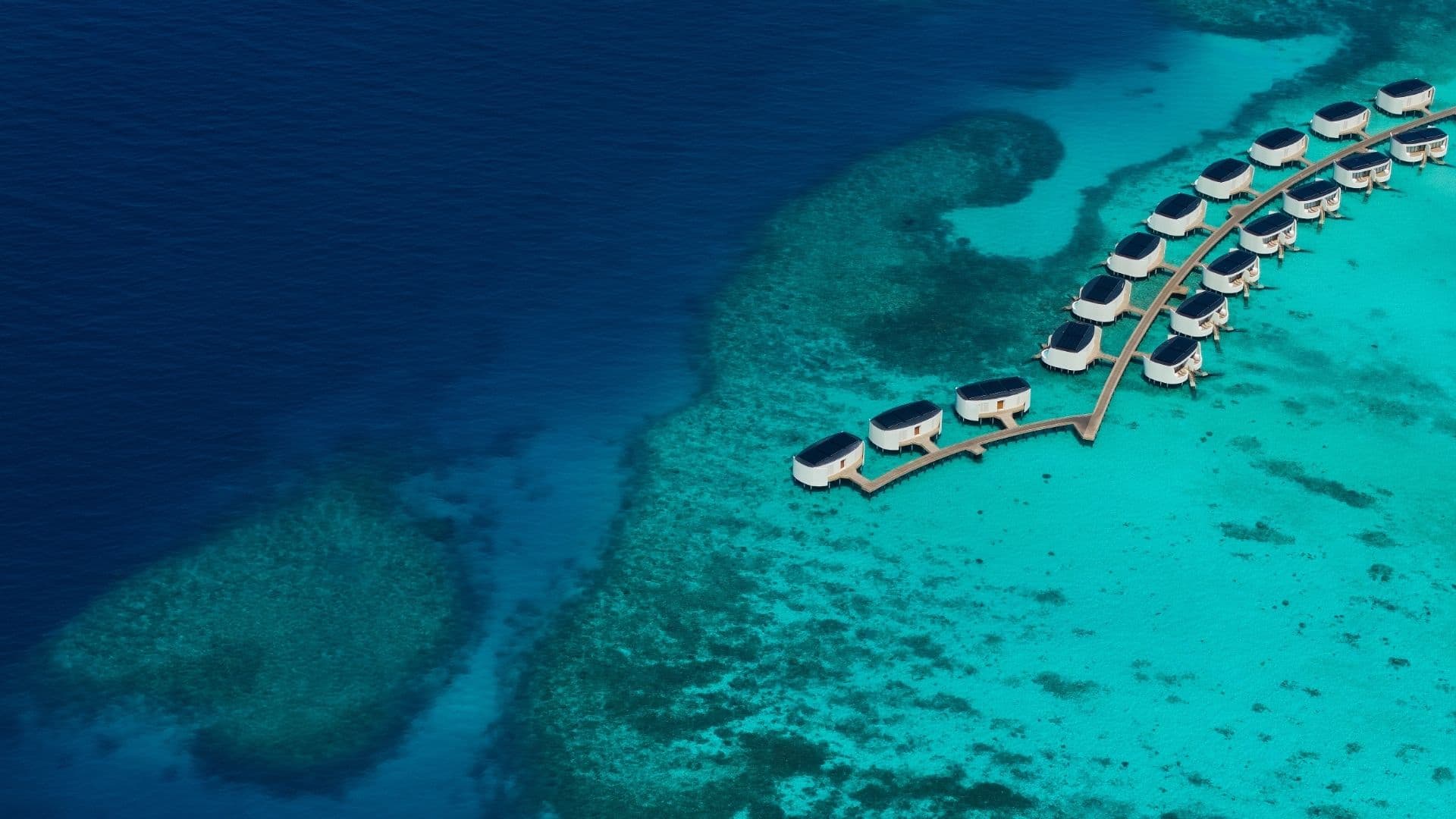 High-altitude aerial shot showing a sweeping overwater boardwalk lined with sleek, rounded modern white overwater villas, extending over shallow turquoise lagoon waters and vibrant coral reef drop-offs into the deep blue ocean.