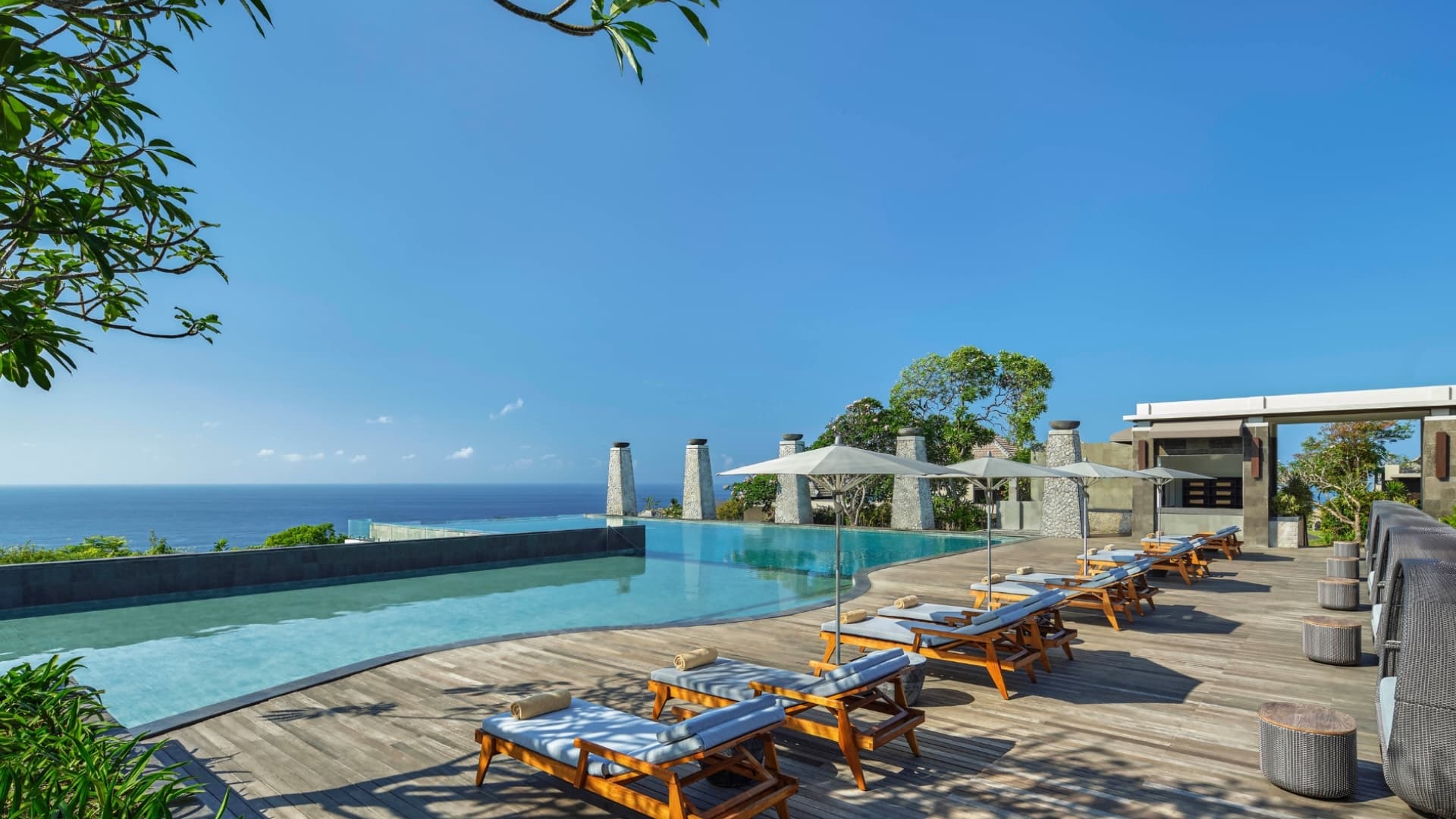 Wide exterior shot of the main infinity pool at Umana Bali featuring wooden sun loungers with white umbrellas lined up along a timber deck, stone pillars, and wide views of the clear blue sky and ocean.